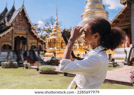 happy black tourist girl taking picture using vintage camera at temple. teenage african american using vintage camera taking temple photo. solo traveler relax doing photo shoot of thailand landmark.