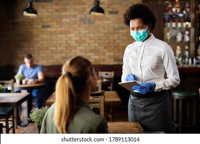 Happy black waitress wearing protective face mask while taking order from customer on a touchpad in a cafe. 