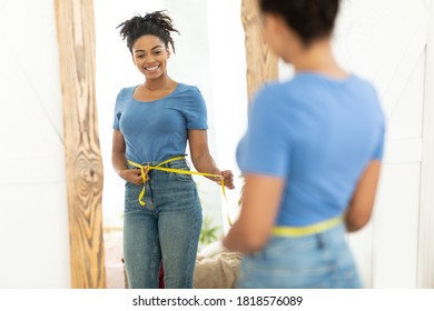 Happy Black Woman After Weight Loss Measuring Thin Waist With Tape Standing Near Mirror At Home. Slimming Concept. Selective Focus