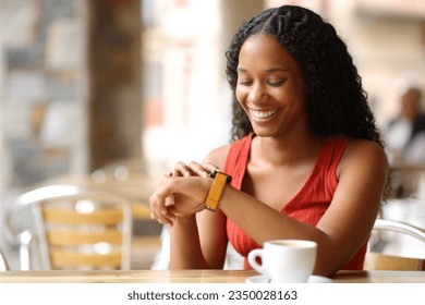 Happy black woman checking smartwatch sitting in a coffee shop terrace - Powered by Shutterstock