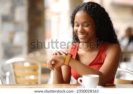 Happy black woman checking smartwatch sitting in a coffee shop terrace