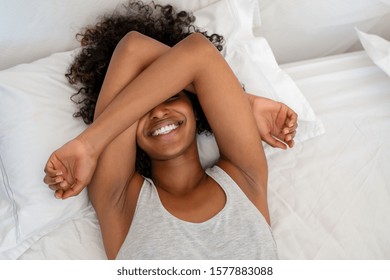 Happy Black Woman Covering Eyes With Arms While Wake Up Happy In The Morning. Top View Of Young African American Woman Waking Up With Happiness And Joy. High Angle View Of Playful Woman Joking On Bed.