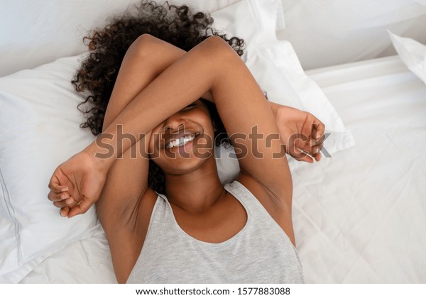 Happy black woman covering eyes with arms while wake\
up happy in the morning. Top view of young african american woman\
waking up with happiness and joy. High angle view of playful woman\
joking on bed.