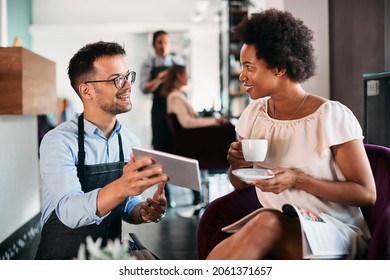 Happy black woman drinking coffee and talking to her hairstylist who is showing her something on touchpad at hair salon. - Powered by Shutterstock