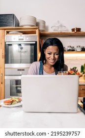 Happy Black Woman Drinking Juice In The Kitchen At Home And Using Computer.