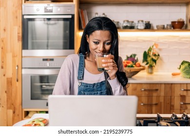 Happy Black Woman Drinking Juice In The Kitchen At Home And Using Computer.