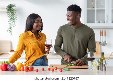 Happy Black Woman Drinking Red Wine While Her Husband Cooking Salad In Kitchen, Young Smiling African American Spouses Having Fun At Home While Preparing Healthy Lunch Together, Copy Space