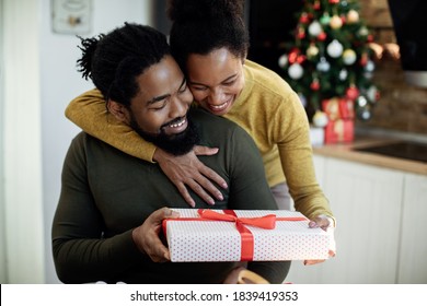 Happy Black Woman Embracing Her Husband And Giving His A Gift On Christmas Day At Home. 