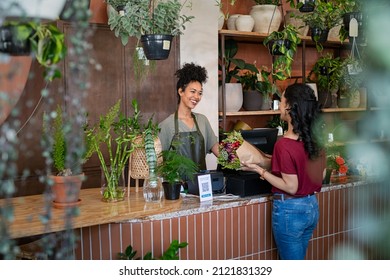 Happy Black Woman Entrepreneur Standing Behind Counter In Plant Store Selling Fresh Flowers To Client. Young Latin Girl Buy A Fresh Bouquet From Florist. Smiling African Woman Botanist Selling Flowers