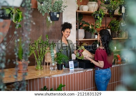 Happy black woman entrepreneur standing behind counter in plant store selling fresh flowers to client. Young latin girl buy a fresh bouquet from florist. Smiling african woman botanist selling flowers