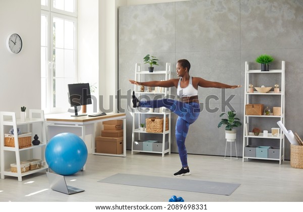 Happy black woman having sports workout at home.\
Active young lady standing in living room, watching online fitness\
workout lesson on modern laptop computer and doing leg swings\
exercise