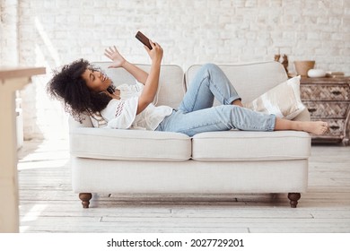 Happy black woman in headphones looking at smartphone, enjoying video meeting with friends and colleagues, online virtual conversation concept. Female student lying on sofa and making video call. - Powered by Shutterstock