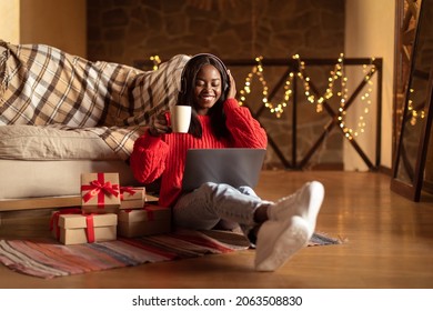 Happy Black Woman In Headphones Using Laptop, Drinking Coffee, Sitting On Floor Near Christmas Gift Boxes At Home, Full Length. Cheery Young Lady Shopping Online, Watching Xmas Movie