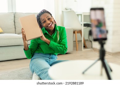 Happy Black Woman Holding Cardboard Box And Talking At Smartphone Camera On Tripod. Unboxing At Home, Online Shopping, Blog, Social Distance And Tech