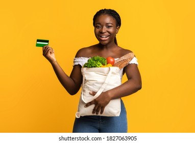 Happy Black Woman Holding Shopping Bag With Groceries And Showing Credit Card, Smiling At Camera, Cheerful African Lady Carrying Package With Fresh Vegetables And Fruits, Isolated On Yellow Background