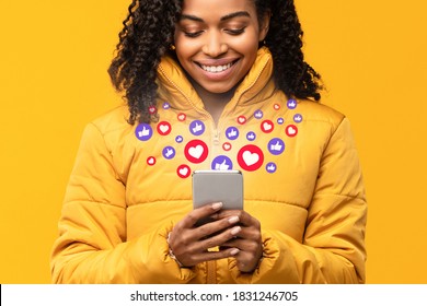 Happy Black Woman Holding Smartphone With Hearts And Like Buttons Standing On Yellow Studio Background. Phone User Networking Online Using Social Media, Reading Feed News, Commenting And Sharing Posts