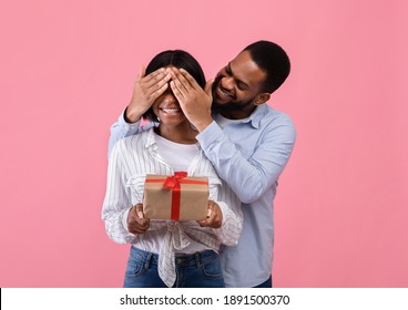 Happy Black Woman Holding Valentine's Gift Box, Loving Boyfriend Covering Her Eyes On Pink Studio Background. Tender Romantic Couple With Holiday Surprise, Giving Festive Present