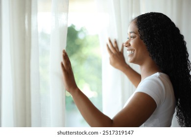 Happy black woman opening curtains and looking through a window at home - Powered by Shutterstock