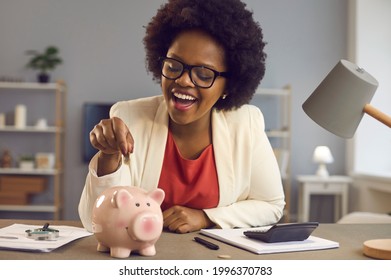 Happy Black Woman Puts Coin In Pink Piggy Bank. Smiling Cheerful Businesswoman Sitting At Office Desk With Calculator And Money Box. People And Finance, Successful Business, Saving Up Concept