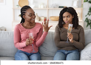 Happy black woman showing her upset and jealous girlfriend engagement ring. Annoyed african american lady cannot share her female friend happiness, drinking champagne together at home