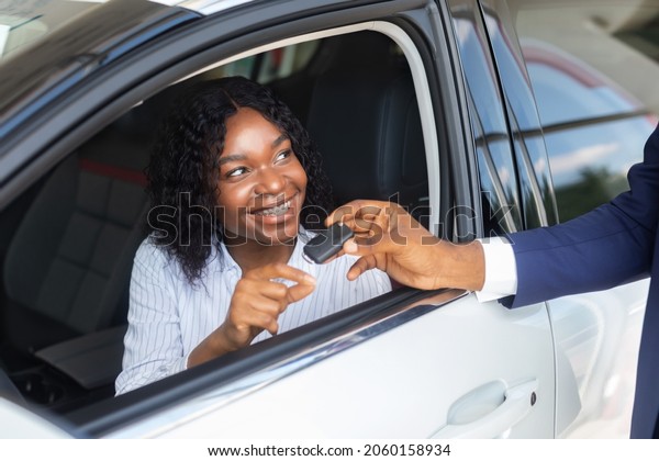 Happy Black Woman Sitting Inside Of Car And Taking\
Key From Salesman After Buying New Automobile In Dealership Center,\
Cheerful African American Female Customer Smiling Through Window,\
Closeup Shot