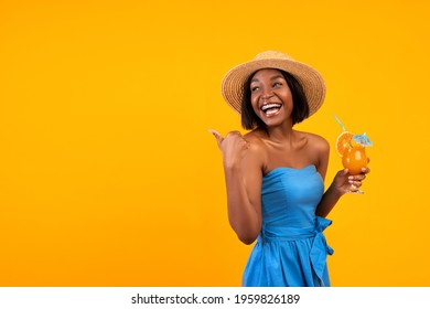 Happy black woman in summer dress and straw hat drinking refreshing tropical cocktail, pointing at empty space on orange studio background. Cheerful lady advertising your summertime product - Powered by Shutterstock