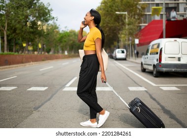 Happy Black Woman Talking On Mobile Phone While Carrying Luggage And Crossing Road