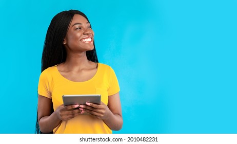 Happy black woman using digital tablet computer looking aside standing over blue studio background. Female browsing internet, looks at copy space. People and gadgets, e-learning. Panorama