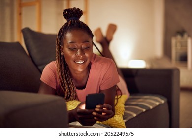 Happy black woman using smart phone and text messaging while relaxing at night in the living room.  - Shutterstock ID 2052249575