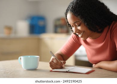 Happy black woman writing in agenda sitting in the kitchen at home - Powered by Shutterstock