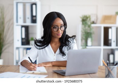 Happy black young business lady working on laptop at office. Focused African American woman taking notes during online work meeting or webinar, writing down important information