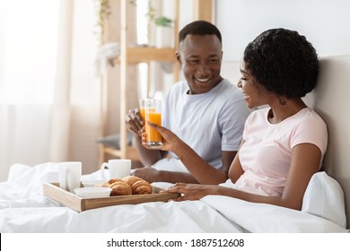 Happy Black Young Couple Sitting In Bed In Morning And Having Breakfast, Cheering With Juice, Side View, Copy Space. Cheerful African American Man And Woman Enjoying Breakfast In Bed, Talking