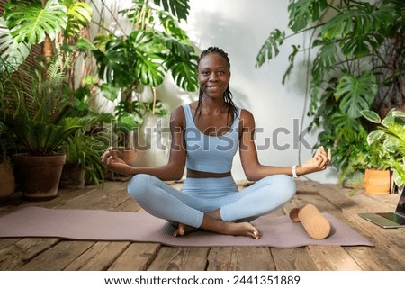 Happy black young female in lotus pose sitting on fit mat looking at camera. Satisfied african american woman meditating in urban jungle place. Home fitness, yoga for healthy lifestyle, stress relieve