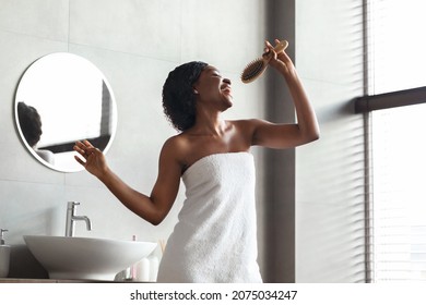 Happy Black Young Woman Covered In Towel Enjoying Haircare, Singing And Dancing With Hairbrush, Having Fun While Brusing Hair After Shower Standing By Window In Bathroom At Home, Copy Space