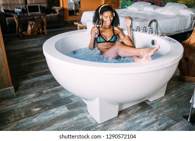 Happy Black Young Woman Relaxing In The Hydro Massage Bath, Drinking Wine And Listen To Music With Headphones