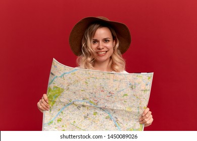 Happy Blond Girl With Curly Hair In A White T-shirt And A Sundown Hat Looks At The Map Looking For Itinerary While Traveling. Concept Of Travel