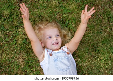 Happy blond girl lying on the green grass, raising his hands up and smiling sincerely. happy childhood.