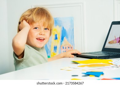 Happy blond little boy using laptop for remote educational videos sitting by the table and turning head