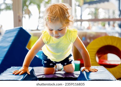 Happy blond little toddler girl having fun and sliding on indoor playground at daycare or nursery. Positive funny baby child smiling. Healthy girl climbing on slide. - Powered by Shutterstock