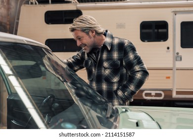 Happy blond man stands by his classic american muscle car in a winter storage. Side view.