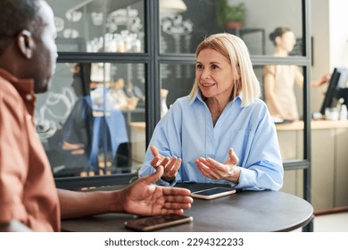 Happy blond mature woman in blue shirt talking to African American male colleague while both sitting by round table in cafe or coffee shop - Powered by Shutterstock