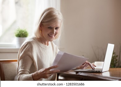 Happy Blond Positive Aged Woman Sitting Alone At Home At Desk Holds Received Document Reading Good News, Notification Paper Checking Bills Or Bank Account Balance Statement Feeling Satisfied And Glad