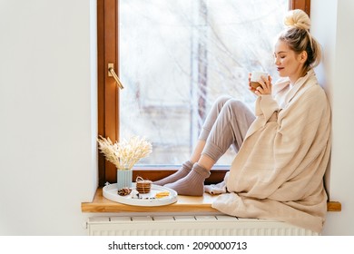 Happy blond woman with bunch hairstyle warming and cover knitted plaid enjoying in her coffee time by the window in cold winter day. Peace of mind and mental health.
