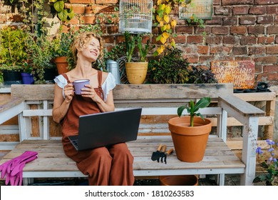 Happy blond woman gardener taking care of houseplants housewife using laptop relaxing on wooden sofa in modern cozy terrace interior, over brick wall. - Powered by Shutterstock