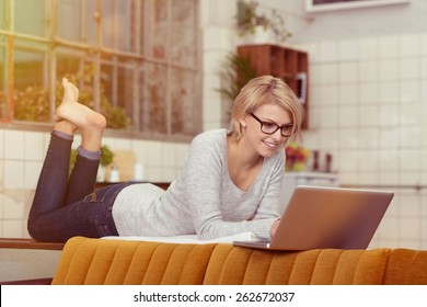 Happy Blond Woman Lying On Her Stomach On The Table Behind The Couch While Using Her Laptop Computer.