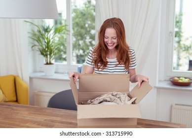 Happy Blond Woman Opening Her Present In A Carton Box On Top Of The Wooden Table Inside The House.