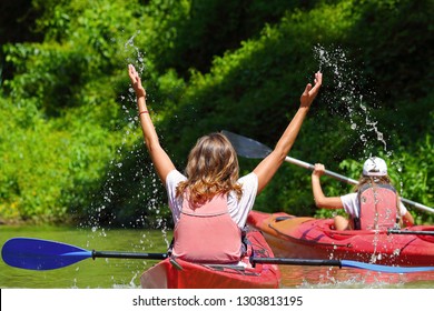 Happy blond woman in red kayak splashing water. Water laughing as she throws it up into the air with her hands. Summer kayaking in the calm Danube river near the shore with green trees.