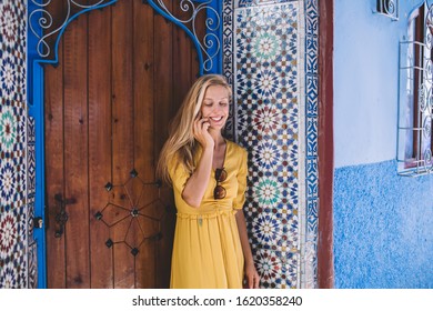 Happy Blond Woman In Trendy Yellow Dress Smiling And Answering Phone Call While Standing Near Entrance Of Traditional House On Street Of Old Town In Morocco