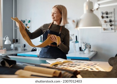 Happy blond woman in workwear looking at piece of suede or leather in her hands while standing by workplace with working supplies - Powered by Shutterstock