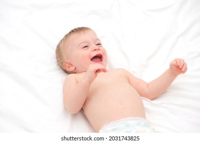 Happy blonde caucasian baby girl about 1 year old laughing lying on back on white bedlinen.Infant having fun before going to bed or after waking up.Cute smiling kid on light bed sheets.Selective focus